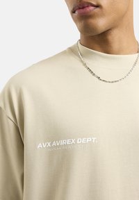 Beżowy sweter z teksturowanym wykończeniem, z białym nadrukowanym napisem na piersi. Okrągły dekolt i widoczny srebrny naszyjnik.