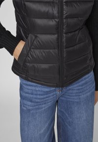 Svart pufferjacka med dragkedja fram, diagonala sömmar och sidofickor. Ribbad stickad ärmar. Kombinerad med blå denimjeans.