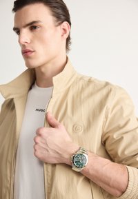 Giacca leggera beige con colletto alto, indossata sopra una t-shirt bianca. Un orologio cronografo verde e argento al polso aggiunge un tocco di dettaglio.