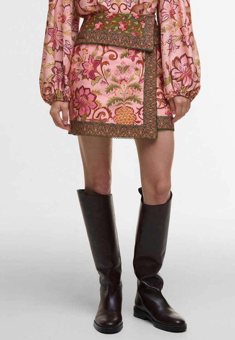 Femme portant une jupe courte rose à motif floral avec une bordure verte, un haut à manches longues assorti, et des bottes en cuir marron foncé montant jusqu'aux genoux.