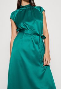 Teal satinklänning med hög krage, ärmkant och knytbart midjebälte. Slät textur med subtil lyster och veckade detaljer vid halsen.