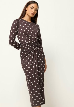 Vrouw draagt een donkebruine midi-jurk met lange mouwen, witte stippen en een gedraaid knoopdetail in de taille, staand tegen een lichte achtergrond.