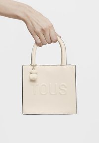 Kremowa skórzana torba noszona na ramię z tłoczonym logo "TOUS", kwadratowy kształt, górne uchwyty oraz odpinany breloczek w kształcie niedźwiadka. Czarne krawędzie kontrastują z korpusem torby.