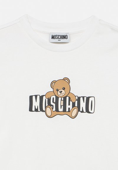 Fehér gyerekpóló, amelyen egy fekete betűkkel írt "MOSCHINO" felirat fölött egy mackó ül.