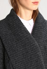 Tmavě šedý pletený cardigan s širokým šálovým límcem, texturovanou látkou a volným střihem, s viditelnými žebrovanými vzory.