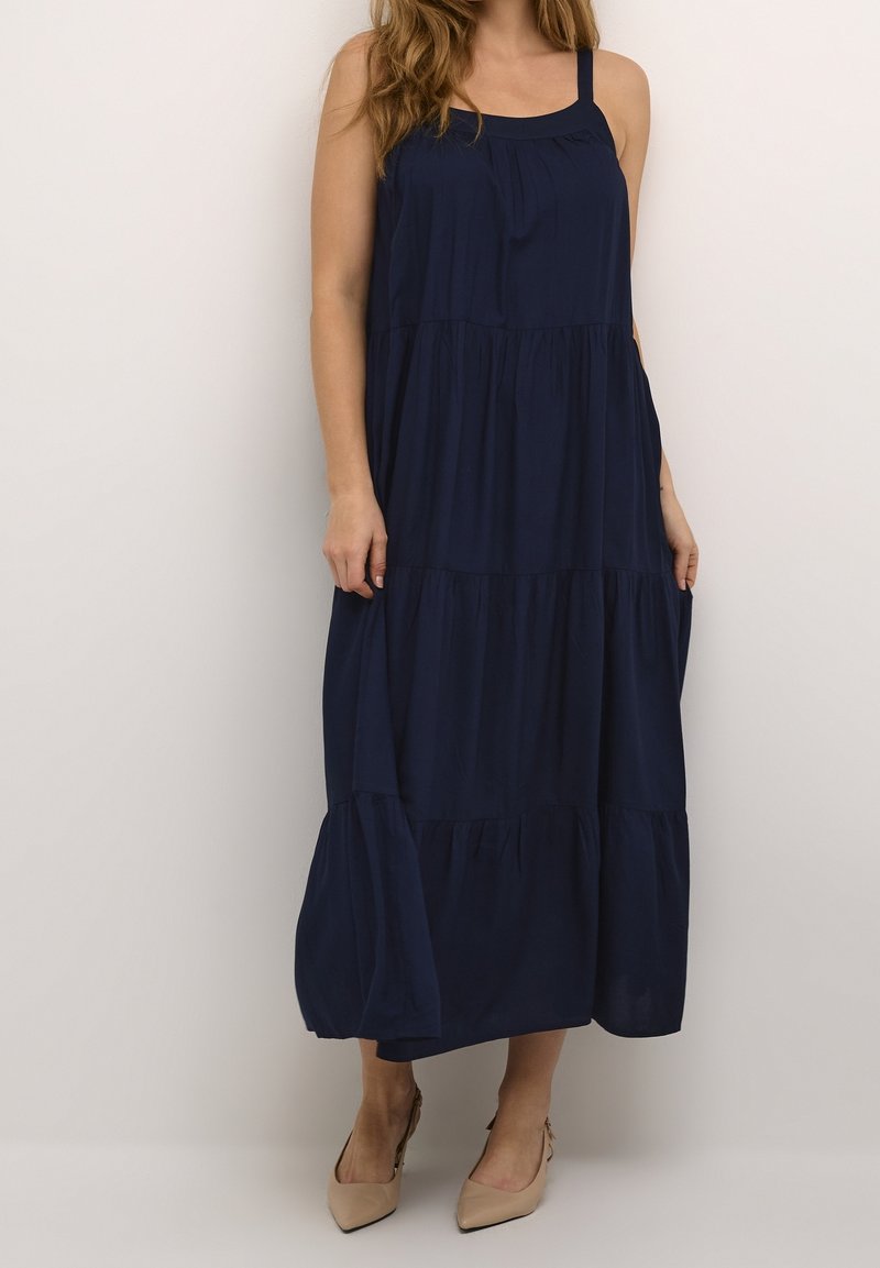 Femme portant une robe longue bleu marine sans manches à plusieurs niveaux avec des talons beiges pointus, debout sur un fond blanc uni.