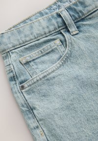 Lichtblauwe spijkerbroek met een voorzak en contrasterende gele stiksels. De stof heeft een zachte textuur met een lichte vervaging.