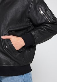 Svart läderbombarjacka med ribbad fåll, sidoficka och dragkedjeficka på ärmen. Slät yta och minimala detaljer i metall.