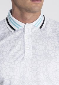 Biela polo tričko má textúrovaný vzor, trojité gombíkovo zapínanie a pruhovaný golier v svetlomodrej a čiernej farbe.