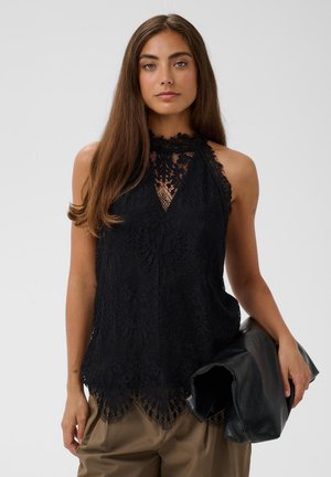 Femme aux longs cheveux bruns portant un top noir en dentelle sans manches et tenant une veste en cuir noire dans sa main droite.
