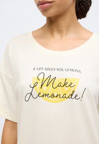 Μπλούζα σε εκρού χρώμα με στρογγυλή λαιμόκοψη που φέρει μαύρο κείμενο, «Αν η ζωή σου δίνει λεμόνια, φτιάξε λεμονάδα!» και ένα κίτρινο γραφικό λεμονιού.