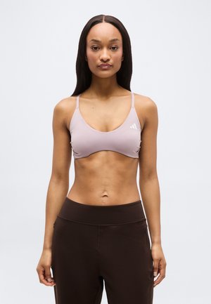 Γυναίκα με μακριά μαλλιά που φοράει ανοιχτό μωβ αθλητικό σουτιέν και σκουρόχρωμο καφέ παντελόνι γυμναστικής, στέκεται μπροστά σε απλό φωτεινό φόντο.