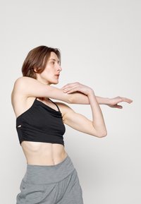 Top cropped atlético preto com detalhes franzidos, combinado com calções de cintura alta cinza claro, usados por um modelo numa pose dinâmica.