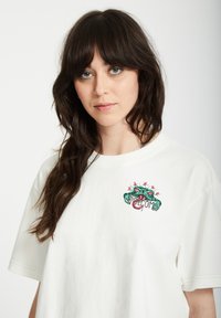 T-shirt en coton blanc avec un design imprimé coloré sur la poitrine gauche représentant un amphibien vert tenant une pancarte sur laquelle est écrit "Bienvenue."
