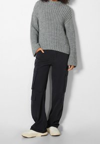 Personne portant un pull en maille épaisse gris, un pantalon cargo noir avec plusieurs poches, et des baskets blanches, debout devant un fond uni.