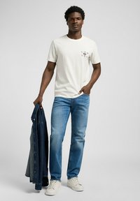 Camiseta blanca de algodón con estampado gráfico, vaqueros de denim azul y una chaqueta de denim oscuro sostenida en la mano izquierda. Zapatillas blancas completan el atuendo.