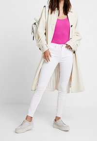 Bílé skinny džíny, růžový žebrovaný top na ramínka a oversized krémový trenčkot. Světle šedé tenisky dotvářejí celý outfit.