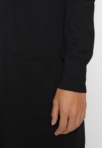 Svart stickad plagg med ribbad textur, långa ärmar och en ficka. Närbild av hand som visar tygets mjukhet och åtsittande ärmmuddar.