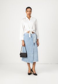 Camicia bianca a maniche lunghe con nodo frontale, gonna avvolgente in denim, borsa nera e scarpe col tacco nere. Tessuti morbidi con dettagli sottili.