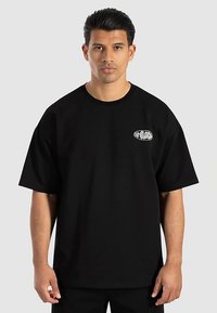 Schwarzes übergroßes T-Shirt aus Baumwolle mit rundem Ausschnitt und kleinem weißen und grünen Logo auf der linken Brustseite.