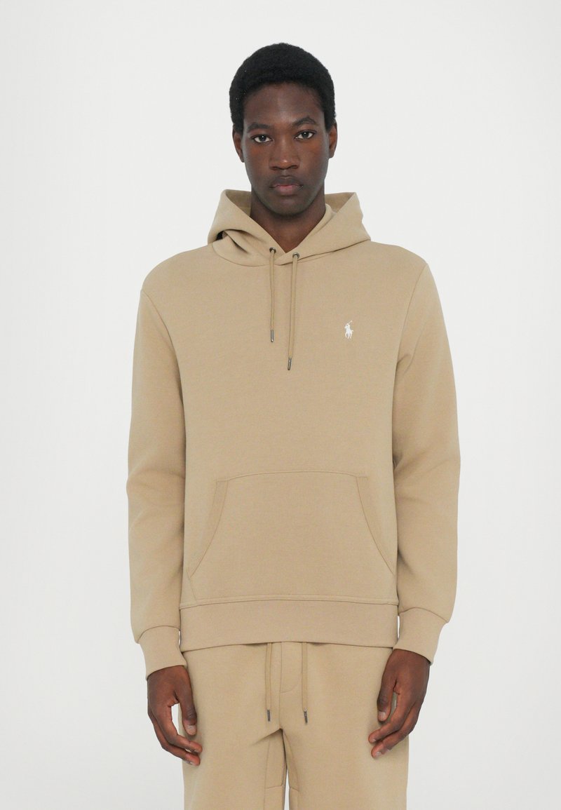 Beige pullover hoodie met een voorzak, verstelbare trekkoordkap en een klein wit logo op de borst. Zachte, gestructureerde stof.