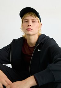 Haut à capuche noir zippé avec un logo blanc, top bordeaux en dessous et casquette noire. Tissu lisse avec une coupe décontractée, position assise contre un mur neutre.