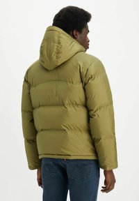 Chaqueta acolchada verde oliva con capucha, con un diseño acolchado y puños elásticos, llevada sobre unos jeans azules. La vista posterior resalta la forma estructurada.