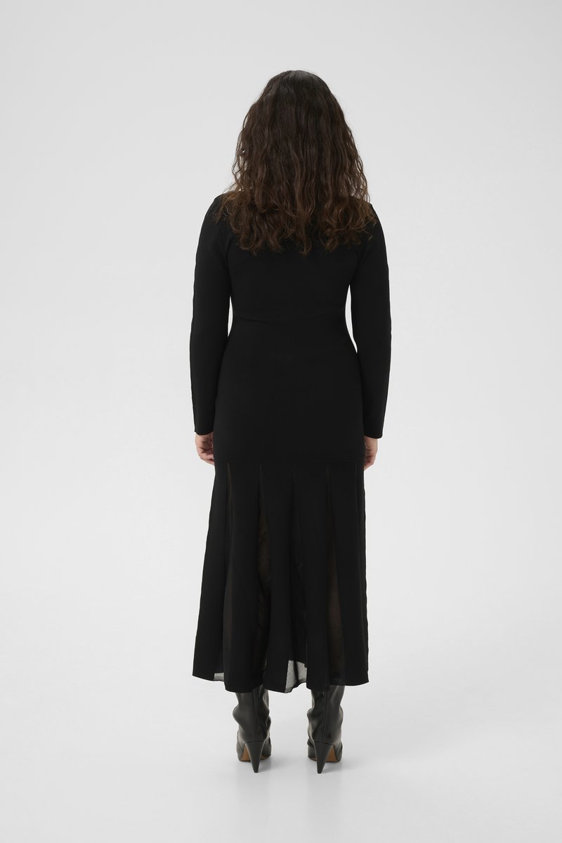 Schwarzes Kleid mit langen Ärmeln, enganliegendem Oberteil und transparenter, gestufter Rock mit seitlichen Schlitzen. Trägt dazu spitze schwarze Stiefel.