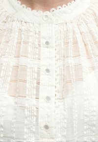 Λευκό μπλουζάκι με δαντελωτή λαιμόκοψη, κουμπιά μπροστά και διάφανο ύφασμα με κάθετες ρίγες και λεπτές κεντητές λεπτομέρειες.