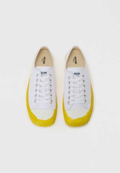 Baltas kanvasu sneakers ar dzelteniem gumijas purngaliem un ārējām zolēm, izceļot sudraba acu caurumus un baltas šņores. Zīmola logo redzams uz mēles.