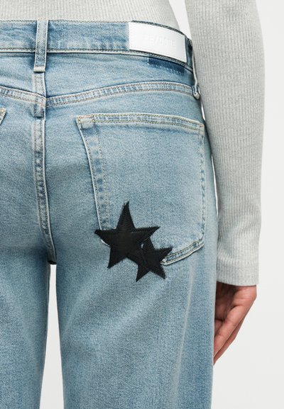 Ανοιχτό μπλε τζιν με μαύρα μπαλώματα σε σχήμα αστεριού στην πίσω τσέπη, φορεμένο με ανοιχτό γκρι ριμπ μακρυμάνικη μπλούζα.