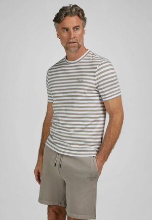 Mann mit grauen Haaren und Bart, der ein grau-weiß gestreiftes T-Shirt und graue Shorts trägt, mit den Händen in den Taschen stehend vor schlichtem Hintergrund.