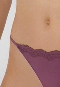 Gros plan d'un torse vêtu de sous-vêtements en dentelle violette avec une fine lanière sur le côté, montrant le nombril et une peau lisse.