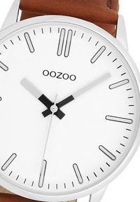 Montre analogique ronde avec un cadran blanc, des index horaires argentés, des aiguilles des heures et des minutes noires, et un bracelet en cuir marron. Nom de la marque "OOZOO" affiché.