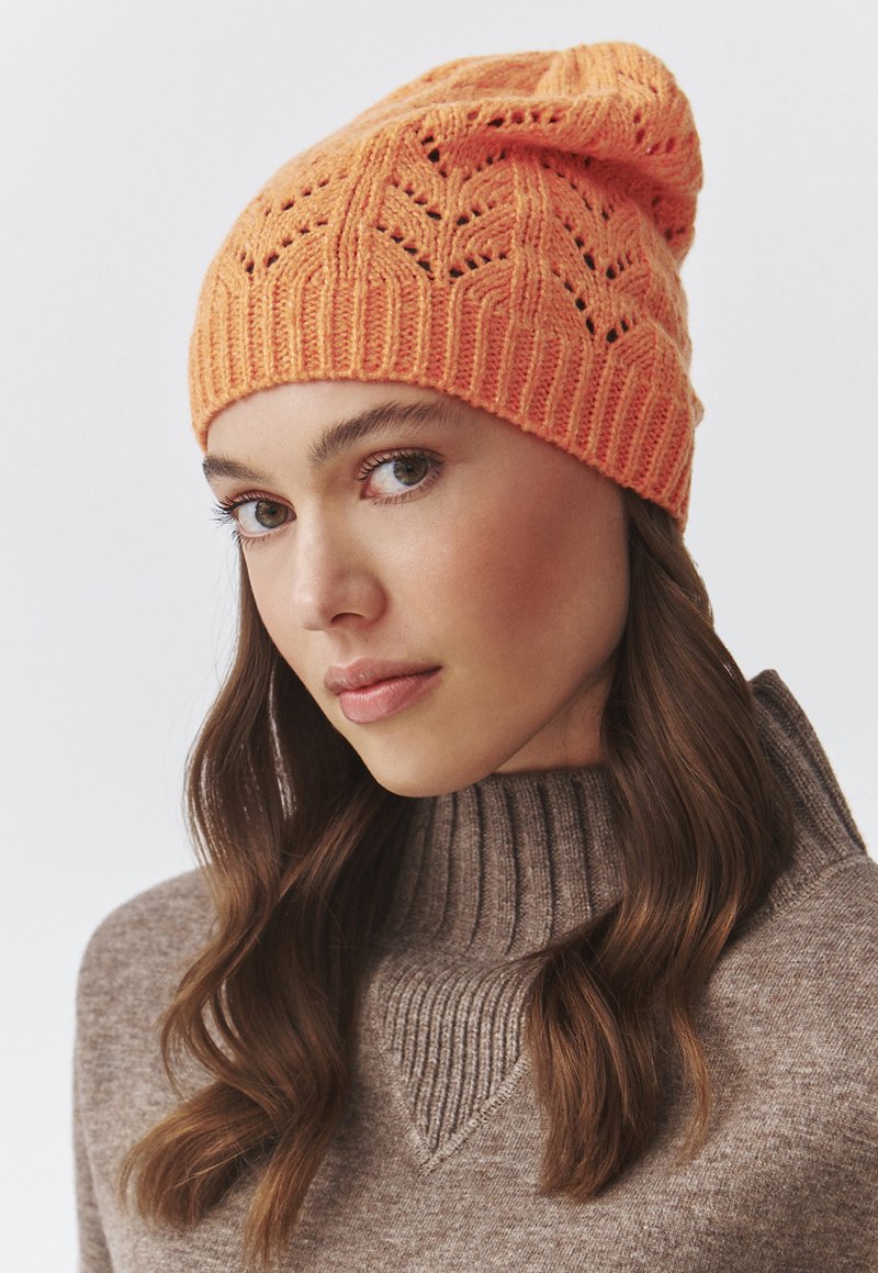 Gorro de punto en naranja brillante con un diseño texturizado que presenta patrones de calado. El ribete es acanalado, lo que proporciona un ajuste ceñido.