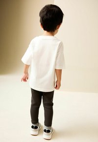 Tricou alb din bumbac, cu mâneci scurte, asortat cu pantaloni negri cu nervuri și adidași albi cu accente negre. Copil văzut din spate.