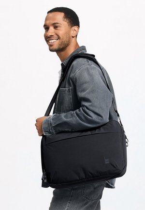 Mann lächelt, trägt eine graue Jeansjacke und trägt eine schwarze Umhängetasche mit dem "GOT BAG"-Logo vor weißem Hintergrund.