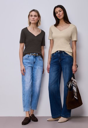 Due donne sono in piedi fianco a fianco, indossano magliette a maniche corte con scollo a V, jeans blu, mocassini marroni e scarpe beige; una di loro tiene in mano una borsa marrone con una sciarpa a motivo.