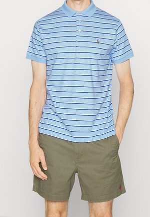 Mann in hellblauem Poloshirt mit dunkelblauen horizontalen Streifen und olivgrünen Shorts, mit den Händen in den Taschen, stehend vor einem einfarbigen Hintergrund.
