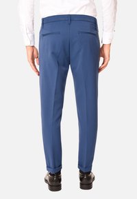 Pantaloni tailored blu in tessuto liscio, slim fit, con due tasche posteriori. Cuffs arrotolati sopra scarpe nere lucide. Camicia bianca visibile nella parte superiore.