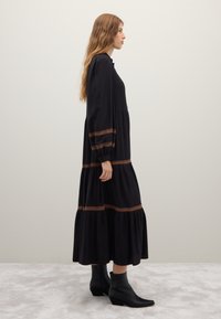 Black long-sleeve dress with a tiered design, featuring contrasting brown trim on sleeves and bodice. Paired with sleek black ankle boots.