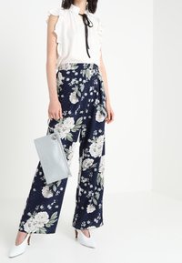 Vit volangblus med svart slips, marinblå blommiga vida byxor, silverhandväska och vita spetsiga klackskor.