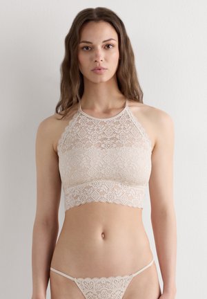 Młoda kobieta z długimi brązowymi włosami, ubrana w beżowy koronkowy biustonosz bralette i pasujące koronkowe stringi, stojąca na tle jednolitego jasnego tła.