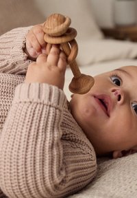 Holzrasselspielzeug, das von einem Baby gehalten wird, mit einer glatten, natürlichen Oberfläche und zylindrischem Design. Das Baby trägt einen strukturierten, hellbeigen Pullover.