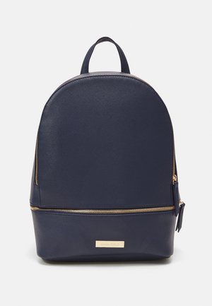 Sac à dos texturé bleu marine avec fermeture éclair et plaque logo dorées, poignée supérieure et compartiment zippé à l'avant, posé sur un fond blanc.