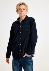 Tmavě modrá manšestrová košile s knoflíkovým zapínáním, kapsou na hrudi a dřevěnými knoflíky. Nošeno s světle modrými džíny, s minimálně viditelnou strukturou.