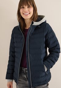 Veste matelassée bleu marine avec capuche grise, texture matelassée, fermeture éclair avant et poches latérales. Portée sur un pull violet foncé.