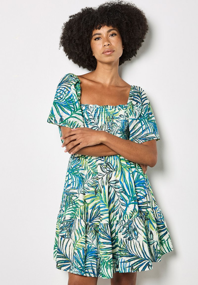 Femme aux cheveux bouclés, bras croisés, portant une robe à manches courtes et col carré avec un motif de feuilles tropicales vertes et bleues, se tenant contre un mur blanc.