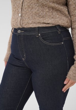 Gros plan d’une personne portant un jean skinny bleu foncé et un pull en tricot marron avec des boutons en perle, main reposant près de la hanche.