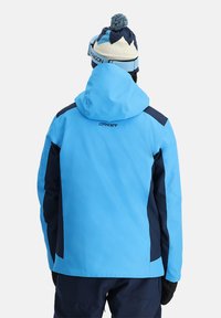 Blaue Skijacke mit marinefarbenen Akzenten, einer Kapuze und einer weichen Textur. Auf der Rückseite befindet sich das Logo "SPYDER" direkt unter dem Kragen.
